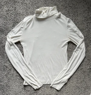 Vit polotopp med långa ärmar - Säljer en stilren vit polotopp med långa ärmar. Toppen har en hög krage och är tillverkad i ett mjukt, stretchigt material som sitter skönt mot kroppen. Perfekt att styla med jeans eller kjol för en clean look.