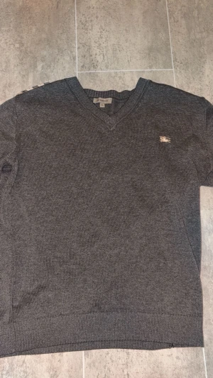 Burberry stickad tröja - Säljer en klassisk grå Burberry-stickad tröja i storlek XL. Stilren modell med v-ringning och ikoniska Burberry-Detaljer på axlarna i det välkända rutmönstret. Liten broderad Burberry-logga på bröstet.