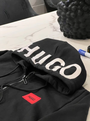 Svart hoodie från Hugo boss - Svart hoodie från Hugo med stor vit logga på huvan och röd patch med Hugo-tryck på bröstet. Tröjan har dragkedja framtill, snörning vid huvan och två fickor. Mjuk bomullskänsla och ribbade muddar vid ärmslut och nederkant. Perfekt för chill och streetwear.