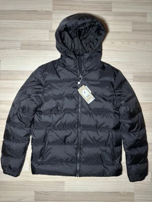 Svart pufferjacka med huva NEO MON DO - Snygg svart pufferjacka från NEO MON DO i storlek M. Jackan har huva, dragkedja framtill och två sidofickor med dragkedja. Tillverkad i polyester och perfekt för kalla dagar. Enkel, stilren design som funkar till allt.