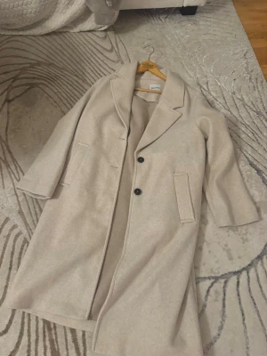Beige kappa från Bershka - Stilren beige kappa från Bershka med klassisk krage och två knappar framtill. Jackan har raka ärmar, två fickor och en slits bak för extra rörelsefrihet. Perfekt för dig som vill ha en enkel men snygg look under kyligare dagar.