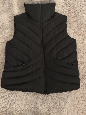 Svart quiltad dunväst Fateh Collection - Säljer en svart quiltad dunväst från Fateh Collection i storlek M. Västen har hög krage, dragkedja framtill och två fickor. Insidan är fodrad med mjukt material och har justerbar dragsko i midjan. Perfekt för lager på lager under kyliga dagar.