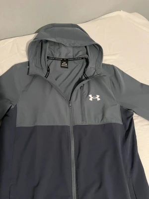 Blå/mörkgrå  vindjacka från Under Armour - tja en riktig skön vindjacka i storlek (S) skicken på den är 10/10! absolut inget fel på den verkan skada eller nån typ av fläck.  Materialet är lätt och känns skönt att ha på sig den💯🙌🏻😄 