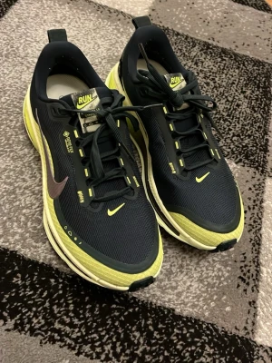 Nike Gore-Tex löparskor svart/gul - Nike löparskor med Gore-Tex material, svarta med gula detaljer och reflexer. Swoosh-logga i lila, robust sula med Storm-Tread för extra grepp. Skorna har snörning och RUN-märke på plösen. Perfekta för löpning i alla väder. Helt ny men har inte kvitto 