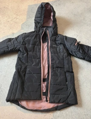 Svart pufferjacka från OTP Sportswear - Säljer en svart pufferjacka från OTP Sportswear med ljusrosa foder och dragkedja. Jackan har huva, snörning i nederkant och praktiska knappar. Perfekt för kyliga dagar och har en snygg, sportig look. OBS!! - Barnjacka som är i storlek - 146. 