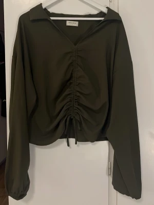 Olivgrön blus med snörning  - Säljer en olivgrön blus med lång ärm och krage. Toppen har snygg snörning framtill som ger en rynkad effekt. Perfekt till jeans eller kjol för en trendig look.