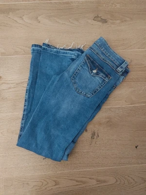 Blå jeans från Gina Tricot, storlek 170 - Säljer ett par klassiska blå jeans från Gina Tricot i storlek 170. Jeansen har bootcut ben med råa, fransiga benslut och femficksmodell. Liten hål vid ena fickan som enkelt går att sy ihop!