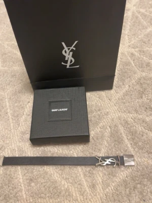 Saint Laurent svart läderarmband - Säljer ett stilrent svart armband från Saint Laurent i läder med silverfärgat YSL-logo och metallspänne. Kommer i original ask och påse, perfekt accessoar för dig som gillar exklusiva detaljer.