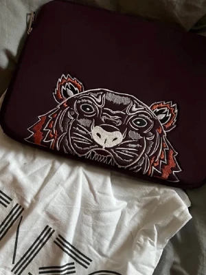 Mörklila datorväska från Kenzo - Säljer en mörklila datorväska från Kenzo med ikoniskt broderat tigerhuvud på ena sidan och Kenzo Paris-logga i rosa på andra sidan. Köpt för 1200kr. Bara fodralet följs med vid köp då jag tappat bort påsen. Har en liten fläck som syns på sista bilden❣️