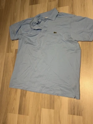 Ljusblå pikétröja från Lacoste - Säljer en klassisk ljusblå pikétröja från Lacoste med den ikoniska krokodilloggan på bröstet. Tröjan har korta ärmar, krage och knappar framtill. Tillverkad i bomull för en skön och luftig känsla.