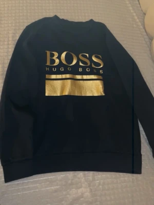  Hugo Boss tröja, mörklblå/guld - Säljer en mörkblå sweatshirt från Hugo Boss med stort guldigt BOSS-tryck och gulddetalj på bröstet. Tröjan har rund halsringning och långa ärmar. Perfekt för dig som vill ha en clean men ändå lyxig look. Storlek M men är lite liten i storleken