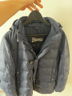 Mörkblå pufferjacka från Herno - Elegant och välarbetad quiltad jacka från Herno, tillverkad i ett lätt men slitstarkt yttertyg med vadderad konstruktion för god värme och komfort. Jackan har en stilren, tidlös design med hög krage, dragkedja och knappstängning som ger både funktion och ett sofistikerat uttryck. Den balanserar perfekt mellan teknisk funktion och klassisk italiensk elegans, vilket gör den lika passande till vardag som till mer uppklädda sammanhang. 