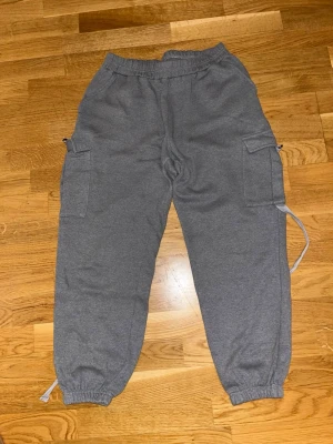 Grå cargopants i mjukis - Säljer ett par grå cargopants i mjukismaterial med resår i midjan och benslut. Byxorna har stora sidofickor och snörning vid anklarna för extra streetkänsla. Perfekta för chill dagar eller när du vill ha en avslappnad look.