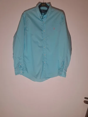 Ljusblå skjorta från Ralph Lauren M - Säljer en ljusblå skjorta från Ralph Lauren med klassisk button-down krage och rosa broderad logga på bröstet. Skjortan har långa ärmar, vita knappar och är i bomull. Perfekt för dig som gillar stilrena plagg med en twist.