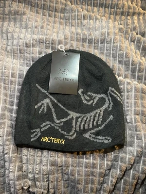 Svart mössa från Arc'teryx - Säljer en svart mössa från Arc'teryx med deras ikoniska fossil-logga i grått och gul broderad text. Mössan är ribbstickad och tillverkad i 100% akryl, perfekt för kalla dagar. Snygg och enkel design som passar till det mesta.