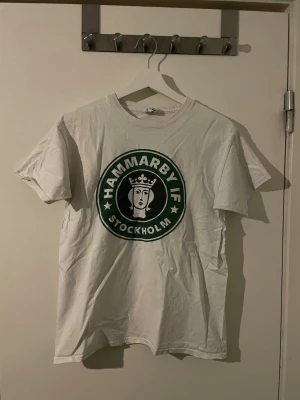 Vit Hammarby IF t-shirt - Vit t-shirt med stort Hammarby IF Stockholm-tryck i grönt och svart på bröstet. Klassisk passform med rund hals och korta ärmar. Perfekt för dig som vill visa ditt stöd för laget på ett stilrent sätt.