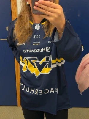 HV71 hockeytröja blå och gul - Säljer en blå HV71 hockeytröja med gula och vita detaljer samt tryck på bröstet och armarna. Oversized passform och långärmad modell, perfekt för dig som vill stötta laget på match 