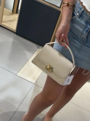 Beige handväska med gulddetaljer - Snygg beige handväska i stilren design med guldfärgat spänne och kedja. Väskan har både ett handtag och en axelrem i kedja. Nypris 200-250 och använd 1 gång 