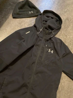 Svart vindjacka från Under Armour - Säljer en svart vindjacka från Under Armour med huva och justerbara snören. Jackan har dragkedja framtill, bröstficka med dragkedja och logga på bröstet. Perfekt för blåsiga dagar och sportiga outfits. Mössan ingår!