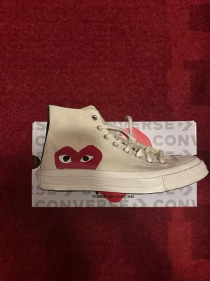 Converse x Comme des Garçons Play - Säljer ett par Converse Chuck Taylor All Star 70s i samarbete med Comme des Garçons Play. Skorna är vita med det ikoniska röda hjärta-ögon-trycket på sidan och klassisk Converse-märke på andra sidan. Hög modell med snörning och canvasmaterial. Storlek 42.5