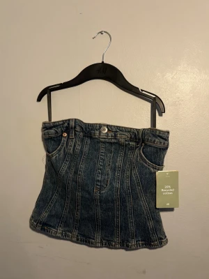 Jeans tubtopp från H&M - Säljer en snygg tubtopp i denim från H&M, gjord av 20% återvunnen bomull. Toppen har klassiska jeanssömmar, bälteshällor och dragkedja i sidan för enkel påklädning. Perfekt för dig som vill ha en edgy och trendig look.