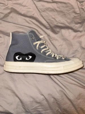Comme des Garçons  x converse high tops - Säljer ett par  Comme des Garçons x Converse i grått tyg med vita snören och sula. Ena sidan har det ikoniska All Star-märket och andra sidan har ett svart hjärta med ögon. Perfekta för dig som vill sticka ut lite extra.