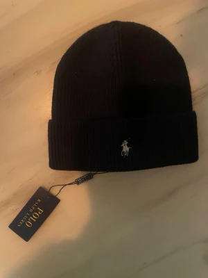 Mörk blå mössa från Polo Ralph Lauren - Säljer en svart ribbstickad mössa från Polo Ralph Lauren med klassisk broderad logga framtill. Mössan är i mjukt material och har en uppvikt kant för extra stilpoäng. Perfekt accessoar för kyliga dagar.