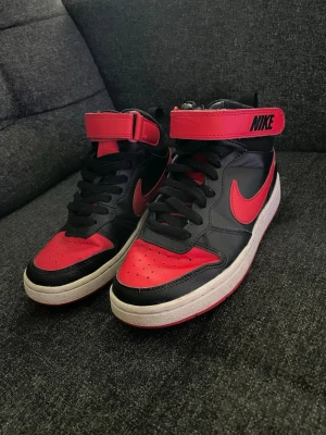 Svarta och röda Nike sneakers - Säljer ett par svarta och röda Nike sneakers med högt skaft och kardborreband upptill. Skorna har klassisk Nike-logga på sidan och bak, svart snörning och vit sula. Materialet är en mix av skinn och textil, och färgkombon ger en riktigt sportig vibe.