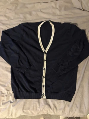 Marinblå kofta från Sand Leisure - Säljer en marinblå kofta från Sand Leisure med kontrasterande vit ribbad kant och knappar framtill. Klassisk v-ringad modell! || Sick: 8/10 || Pris: 599 || Storlek: M ||