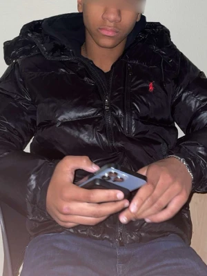 Svart pufferjacka från Polo Ralph Lauren - Säljer en svart pufferjacka från Polo Ralph Lauren med röd broderad logga på bröstet. Jackan har huva, dragkedja framtill och är tillverkad i glansigt syntetmaterial. Perfekt för kalla dagar och riktigt snygg streetstil.