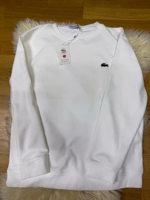 Lacoste sweatshirt, HELT NY!!!! - HELT NY!!! Säljer en clean vit sweatshirt från Lacoste med klassisk rund hals och den ikoniska krokodilloggan broderad på bröstet. Tröjan har långa ärmar och ribbade muddar vid ärmslut och nederkant. Perfekt för en stilren och enkel look.