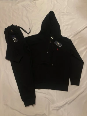 Svart Polo Ralph lauren set - Svart hoodie samt byxor från Polo Ralph Lauren med dragkedja framtill och huva. Hoodien har en röd broderad logga på bröstet och är tillverkad i mjuk bomull. Perfekt för en chill och stilren look.