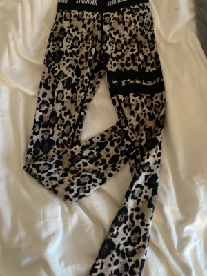 Leopardmönstrade leggings från Stronger - Snygga leggings från Stronger med leopardmönster i beige, svart och bruna toner. Hög midja med elastisk linning där det står 'STRONGER' och svarta detaljer på benen. Perfekta för träning eller chill.