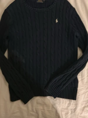 Mörkblå kabelstickad tröja Polo Ralph Lauren - Kabelstickad mörkblå tröja från Polo Ralph Lauren med klassisk rund halsringning och den ikoniska vita logon broderad på bröstet. Tröjan har långa ärmar och ribbade muddar vid ärmslut och nederkant. Perfekt för lager på lager under höst och vinter. Den har en vit fläck men syns knappt i vanligt ljus bara med kamera. 
