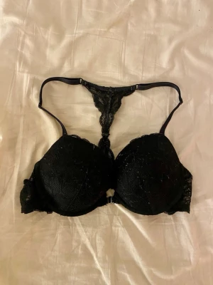 Svart spets-bh från Lindex Tease - Säljer en svart bh från Lindex Tease med snygga spetsdetaljer och unik ryggdesign. Bh:n har vadderade kupor och tunna axelband som går ihop i en spetsig T-form baktill. 