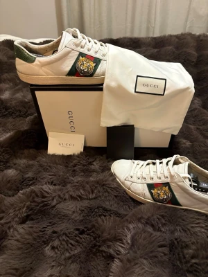 Gucci Ace sneakers med tigerpatch - Gucci Ace sneakers i vitt skinn med klassiska gröna och röda ränder på sidan och broderad tigerpatch. Baksidan har en grön och en röd detalj i ormskinnslook. Skorna har snörning och platt sula med Gucci-logga undertill. Kommer med originalkartong och dustbag.