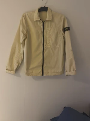 Beige vindjacka från Stone Island - Säljer en beige vindjacka från Stone Island. Jackan har en klassisk krage, svart dragkedja framtill, bröstficka och den ikoniska loggan på ärmen.  Jackan har en mindre fläck på höger arm vilket jag kommer ta på vid köp. Skick är på 8/10, hör av er vid funderingar! Den är i storlek juniors men passar mer åt S hållet. 