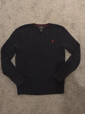 Blå  stickad tröja från Ralph Lauren - Kabelstickad mörkblå tröja från Polo Ralph Lauren med klassisk röd broderad logga på bröstet. Tröjan har rund halsringning, ribbade muddar och är långärmad. Perfekt för dig som gillar stilrena och tidlösa plagg. Tröjan är i nytt skick då den inte passar mig längre och använt den fåtal gånger tröjan är i junior ålder 14/16 år men passar xs. Köpt på NK för 1600 kr 