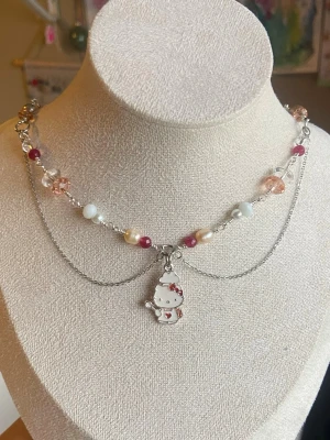Hello Kitty pärlchoker med kedja - Choker med pärlor i vitt, rosa och beige samt silverfärgad kedja. Hängsmycke med Hello Kitty i mitten, dekorerad med röda detaljer. Pärlorna är blandade i form och färg, och kedjan ger ett lager-på-lager utseende. Perfekt för dig som gillar söta accessoarer.