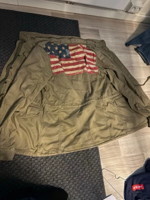 Olivgrön parkajacka Ralph Lauren - Säljer en olivgrön parkajacka från Denim & Supply Ralph Lauren. Jackan har flera fickor, dragkedja och knappar framtill samt en stor amerikansk flagga på ryggen. Tillverkad i bomull och har justerbar midja med snörning för skön passform. Denna jacka går ej att få tag i längre i butik och just denna modell  är väldigt sällsynt därför kommer jag lägga priset högt men går att diskutera 