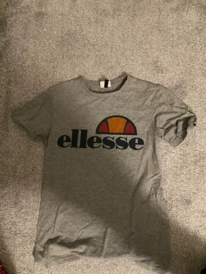 Grå ellesse t-shirt med logga - Säljer en grå t-shirt från ellesse med stor tryckt logga i svart, orange och rött på bröstet. Klassisk rund hals och korta ärmar. Tillverkad i mjuk bomull som är skön att ha på sig. Perfekt för en avslappnad och sportig stil. Köpt för 300 kr säljs för 80 kr!