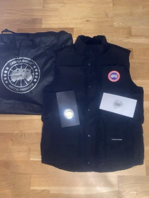 Svart dunväst från Canada Goose - Säljer en svart dunväst från Canada Goose med klassisk logga på bröstet och hög krage. Västen har tryckknappar framtill och två stora fickor. Materialet är slitstarkt och fyllningen består av dun, perfekt för kyliga dagar. Kommer med originalpåse och certifikat. 😊