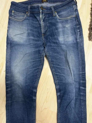 Blå Lee Malone jeans W33 L32 - Snygga blå jeans från Lee i modellen Malone. Jeansen har klassisk femficksdesign, normal passform och raka ben. Färgen är mellanblå med slitningar och snygg tvätt. Perfekta för en avslappnad stil och funkar året runt.