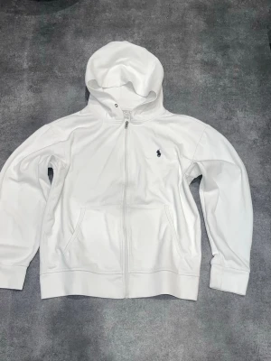 RL Zip Hoodie - Vit zip hoodie från Polo Ralph Lauren. Riktigt clean och skön modell som funkar till typ allt. Hel dragkedja, fickor fram och broderad Polo-logga på   Det saknas en öljett på vänster sida (se bild 3). I övrigt bra skick och redo att användas direkt. Perfekt om du vill ha en äkta Polo-hoodie men inte betala fullpris.