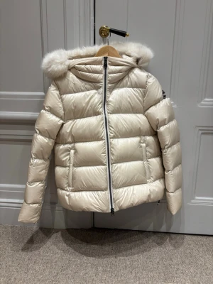 Beige dunjacka med fuskpäls - Snygg beige dunjacka med huva och fuskpälsdetalj. Jackan har quiltad design, dragkedja framtill och två fickor. Perfekt för kalla dagar och ger en riktigt trendig look.