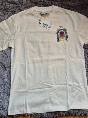 Vit t-shirt från Casablanca med tryck - Säljer en vit t-shirt från Casablanca i storlek L. T-shirten har ett färgstarkt tryck med motiv av en byggnad och gröna blad både fram och bak, samt texten 'Avenida Casablanca'. Klassisk passform och rund hals. Tillverkad i mjuk bomull.