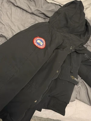 Svart dunjacka från Canada Goose - Säljer en svart dunjacka från Canada Goose med huva och klassisk patch på ärmen. Jackan har dragkedja, knappar och flera fickor, samt ribbade muddar vid ärmslut och nederkant. Perfekt för kalla dagar och riktigt snygg streetstil.