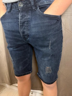 Mörkblå jeansshorts med slitningar - Snygga mörkblå jeansshorts med slitna detaljer och upprullade benslut. Klassisk femficksmodell med knapp och dragkedja framtill. Perfekta för en avslappnad stil och passar till det mesta. Materialet är mjukt denim som ger en skön känsla.