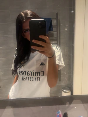 Vit Real Madrid t-shirt Adidas - Säljer en vit Real Madrid t-shirt från Adidas med klubbmärke och sponsortryck på bröstet. Klassisk v-ringning och korta ärmar, perfekt för fotboll eller att bara chilla i. Ikoniska Adidas-ränder på axlarna.