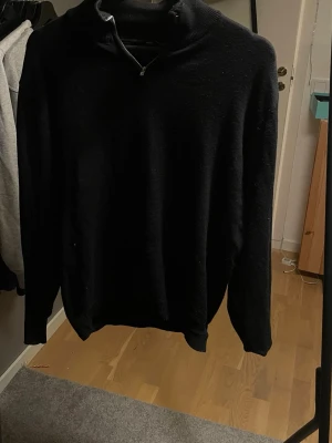 Svart stickad tröja med half zip - En svart stickad tröja med halv dragkedja i halsen. Tröjan har lång ärm och ribbade muddar vid ärmslut och nederkant. Perfekt för kyliga dagar och enkel att matcha med jeans eller byxor.